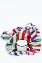 Old metal cup of hot cocoa with marshmallows and a soft colorful knitted scarf on a fluffy the snow
