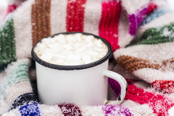 Obraz na płótnie Canvas Old metal cup of hot cocoa with marshmallows and a soft colorful knitted scarf on a fluffy the snow close-up
