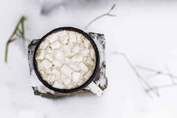 Old metal cup of hot cocoa with marshmallows stands on covered with snow pole outdoors