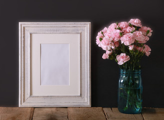 A horizontal presentation of a rustic white wood frame and a teal glass vase with pink carnations.  A black background provides the perfect mock up presentation for your business concept.
