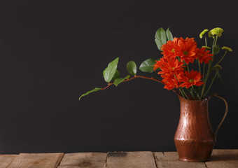 Bright red flowers with green leaves in a copper pitcher vase on a wood table with a striking black background. © cpdprints