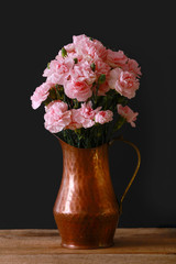 A vertical presentation of a copper vase with a bouquet of pink carnations on a black background. 