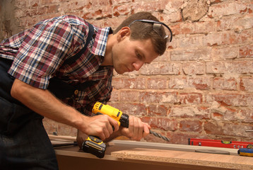 Strong young man wearing worker overalls with hand-operated drill in a carpenter shop (retro style)