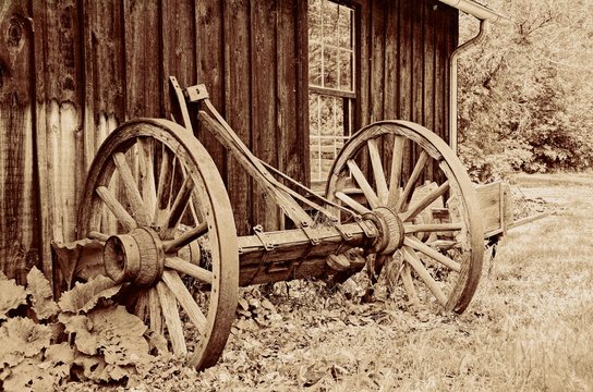 Sepia vintage wagon wheel and axel leaning against a barn