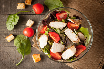 Fresh salad made of tomato, ruccola, chicken breast, arugula, crackers and spices. Caesar salad in a white, transparent bowl on wooden background