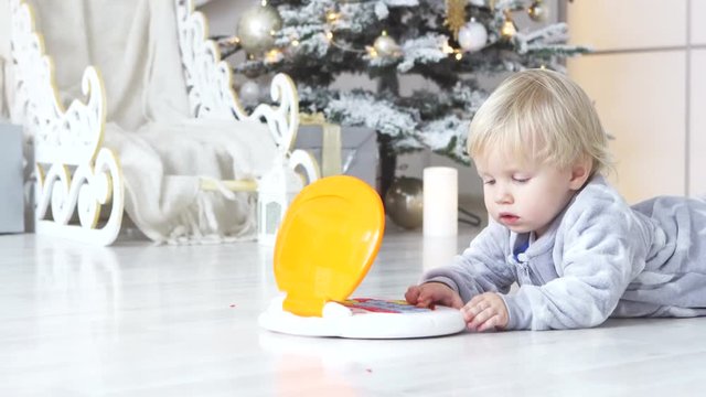 baby in Santa pajamas with stars sits near a Christmas tree with a gift and play with toy tablet. New Year concept with toddler