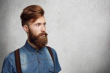 Casual young man with stylish fuzzy beard dressed in denim shirt and suspenders looking ahead of him at blank wall with copy space for your text or promotional content. Isolated shot, horizontal
