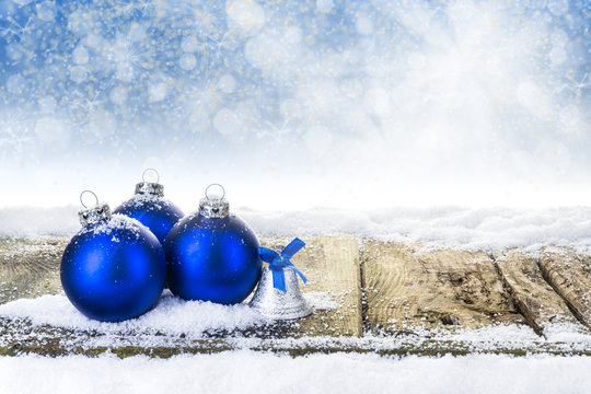 Blaue Weihnachtskugeln und Weihnachtsglocke auf verschneiten Holzbrettern vor Bokehhintergrund