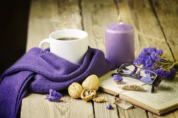 Obraz na płótnie Canvas Cup of coffee wrapped in purple cozy scarf placed with vintage book,walnuts,glasses, dry flowers and purple candle on rustic wooden table.