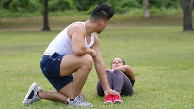 A young Asian man assists his Latina workout buddy while she does her sit-ups.