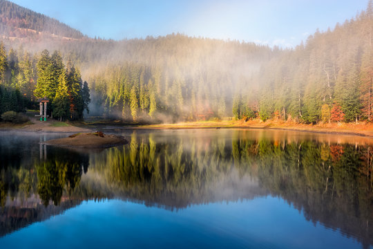 view on crystal clear lake with smoke and reflection on the water near the spruce forest in fog at the foot of the mountain at sunrise