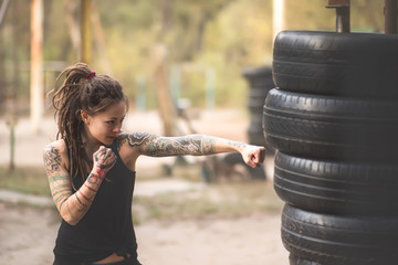 Woman boxer with tatto arms at workout. Young woman is boxing punching bag from car tires. At the head of the girl dreadlocks. Toned photo.
