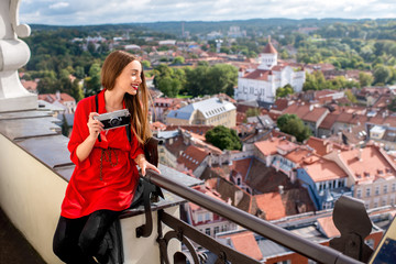 Young female tourist with photo camera enjoying great view on the old town from the tower in Vilnius. Woman having happy vacations in lithuania - obrazy, fototapety, plakaty