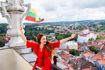 Young female tourist with photo camera and flag enjoying great view on the old town on the tower in Vilnius. Woman having happy vacations in lithuania - obrazy, fototapety, plakaty