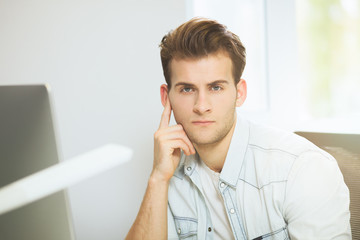 Fototapeta premium A young programmer is looking at the camera. The graphic designer is thinking about the future projects. The young guy is sitting at the computer in the office with the windows behind his back