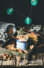 Mug of hot chocolate with whipped cream, cocoa powder, cinnamon, gingerbread cookies, nuts in wooden tray, dark wall with Christmas decoration garland of blue balls at background, selective focus