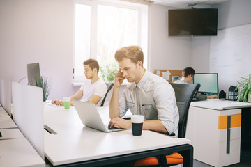 A young programmer is looking at the computer. The graphic designer is thinking about the future projects. The young guy is sitting at the computer in the office with the windows behind his back