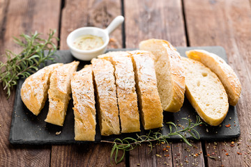 fresh homemade ciabatta bread sliced on stone slate board, olive oil and rosemary on wooden background