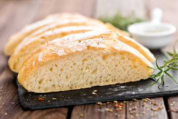 fresh homemade ciabatta bread sliced on stone slate board, olive oil and rosemary on wooden background