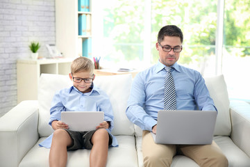 Little son with tablet and father with laptop sitting on sofa at home