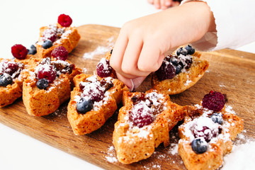Close-up of child hand decorating berry cakes with sugar powder. Young confectioner cooking sweet cookies. Homemade bakery, children culinary, pastry making concept