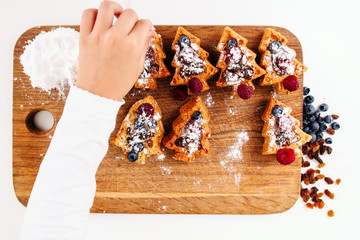Kids hand sprinkle cookies with sugar powder, confectioner pov. Top view on board with set of berry...