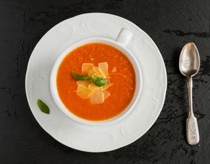 Tomato cream soup with parmesan cheese in white bowl on black stone background. Fresh basil leaves and vegetables, top view.