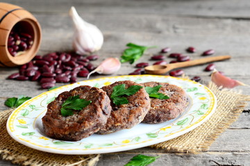 Cutlets made from boiled and mashed red beans on a plate. Scattered uncooked red beans, garlic, fresh parsley, small wooden spoon and decorative barrel on old wooden background. Vintage style. Closeup