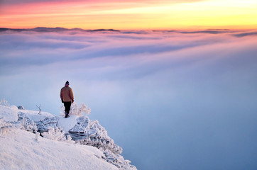 Man standing on the snowy frozen rocks and looking to the valley full of blue dark clouds and beautiful winter sunrise full of vivid colors.