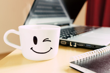 Smiling cup, notepad and laptop on the desk