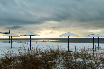 Fototapeta na wymiar Deserted winter snow covered beach with sunset, skyline, snow, snow covered beach umbrellas, dried grass and beautiful sun rays.