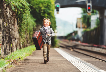 Outdoor portrait of cute little kid boy, run to the train station, wearing beige jacket, trousers, scarf and brown retro shoes, holding old vintage suitcase, small tourist going to vacation