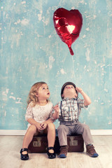 zwei kleine Kinder mit roten Herzluftballon auf alten Koffer