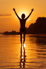 Fototapeta premium Young woman with hands raised standing in water in the sunlight, sunset or sunrise