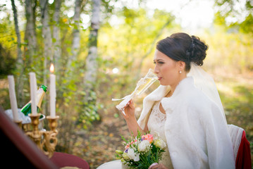 Bride is holding a wedding bouquet and a glass of champagne. Hands newlyweds on the table. 
