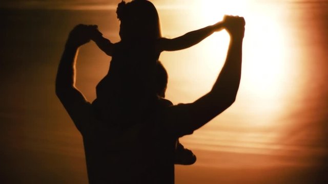 Medium shot of silhouette of unrecognizable man turning around with toddler girl sitting on his shoulders 