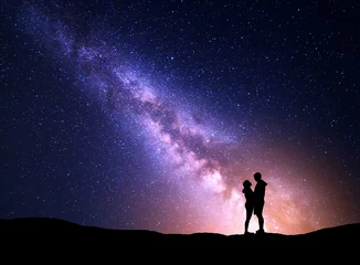Afwasbaar fotobehang Milky Way with silhouette of people. Landscape with night starry sky. Standing man and woman on the mountain with yellow light. Hugging couple against purple milky way. Beautiful galaxy. Universe   © den-belitsky