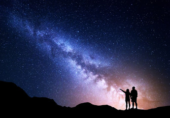 Melkweg met mensen op de berg. Landschap met nachtelijke hemel met sterren en silhouet van staande gelukkige man en vrouw die met de vinger in de sterrenhemel wijzen. Melkweg met reizigers. Prachtig sterrenstelsel