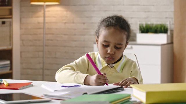 African girl sitting at the table and drawing on paper with pink felt tip pen