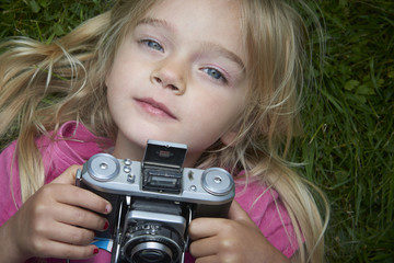 Portrait of little girl taking picture using vintage old retro film camera, lying on grass background