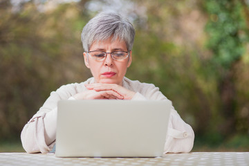 Older woman with gray hair in a park with a gray computer sits on a chair in the autumn outdoor ambience. Shallow depth of field.