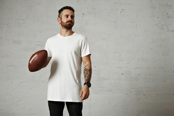 Tattooed and bearded model in plain white shortsleeve t-shirt holding a leather football against gray wall background