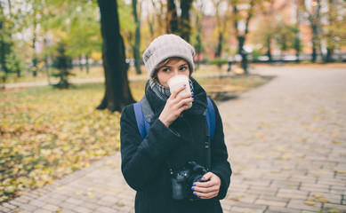 Fototapeta premium Woman in coat holding a cup of coffee latte with milk. Lonely woman stands on a snowy autumn deserted street in the park.