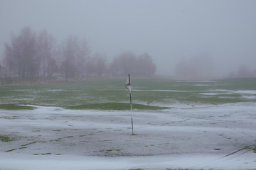 Danish golf course covered in snow, with a flag on a black and white striped pole standing in a hole, on a foggy winter day