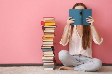Woman sitting on a floor and holding book in front of face on pink background - obrazy, fototapety, plakaty