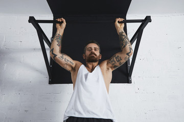 Strong tattooed in white unlabeled tank t-shirt male athlete shows calisthenic moves close up Pullup hold on medium move on pull bar