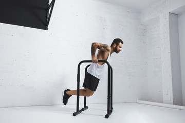 Strong tattooed in white unlabeled tank t-shirt male athlete shows calisthenic moves Holding dip position on parallel bars