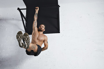 Strong tattooed in white unlabeled tank t-shirt male athlete shows calisthenic moves Hanging on pull bar one arm leg raises or l-sit hold