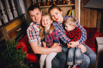Young family is photographed for Christmas card. Mother, father and little daughter with smile look in camera. They recline on a big bed with bright pillows. Room is decorated with Christmas garlands.