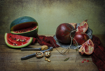 Classic still life with watermelon and pomegranate placed  in vintage silver plate with red silky scarf , some walnut and nut crusher on rustic wooden table.